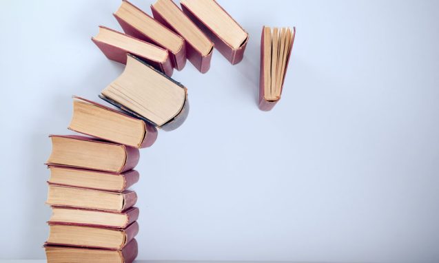 a stack of thirteen books with the top seven beginning to topple over onto a white surface