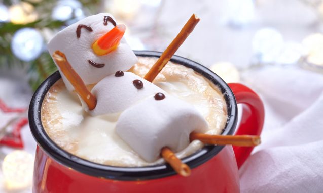 cute marshmallow man lays in a cup of hot cocoa on a table decorated for Christmas