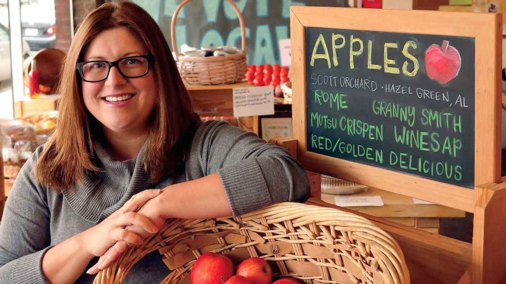A woman in front of a sign listing different types of apples