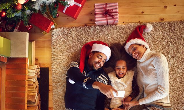 family of three snuggle on a rug to take a photo in front of a fire place under their Christmas tree