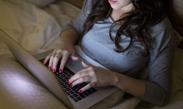 girl with red fingernails types on a laptop in bed