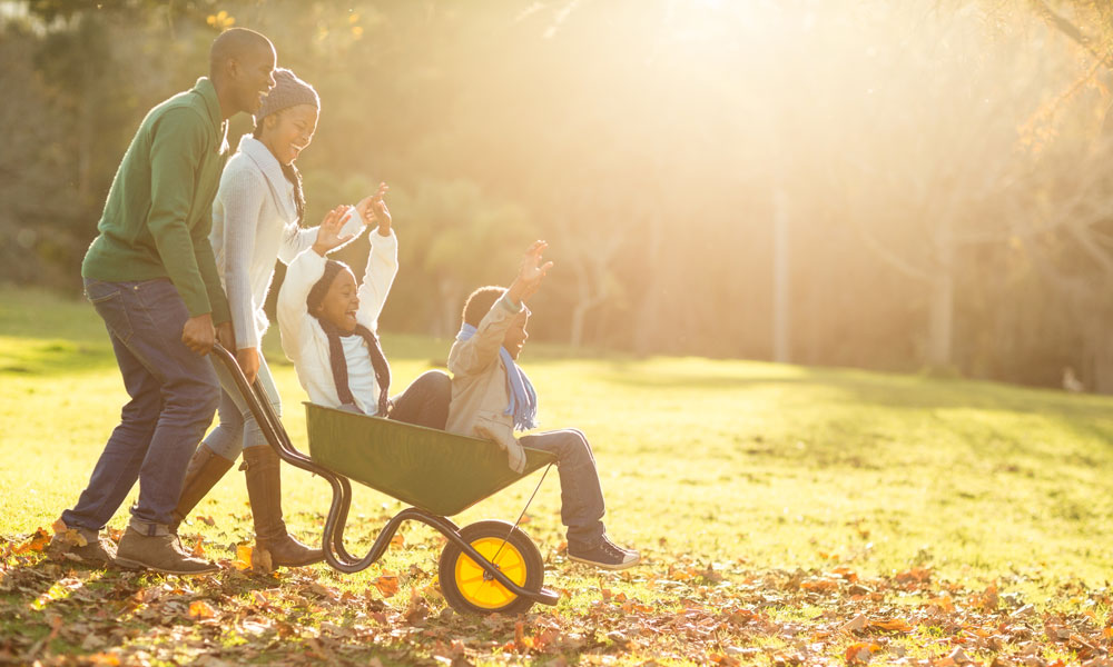 father pushes two young children in a wheelbarrow with wife laughing at his side on a sunny fall day in the park