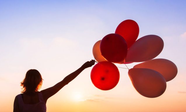 woman holds a cluster of red balloons in front of a sunset