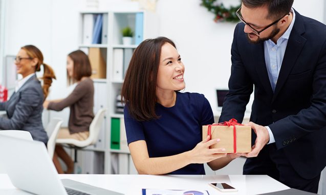 A man hands a boxed gift tied with red ribbon to a woman seated at a desk