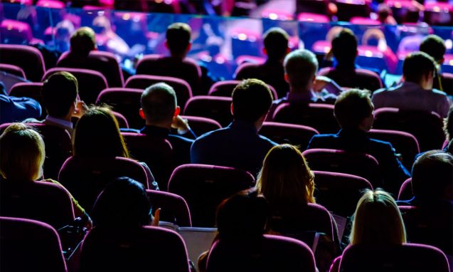 audience members pay attention during a tech conference in a theatre with colored lights on stage