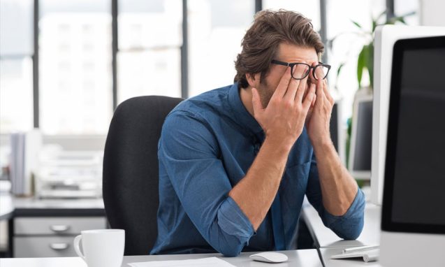 man rubs his face in frustration at his office in front of his desktop
