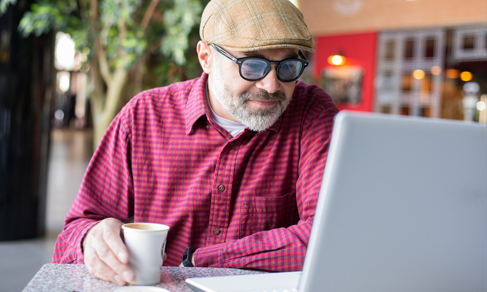 Bearded man wearing eyeglasses and cap holds a cup of coffee while looking at a laptop screen