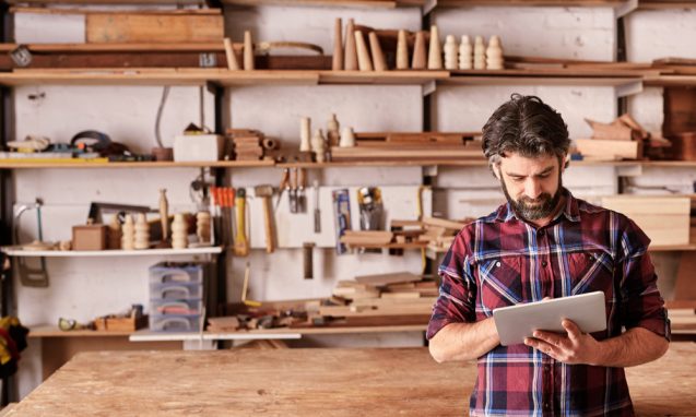 bearded man stands holding an ipad in an artisan wood working shop