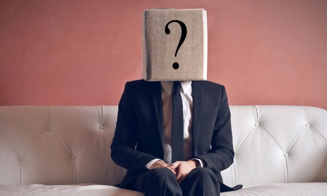 Businessman sits on a cream sofa with a box on his head with a big question mark on it